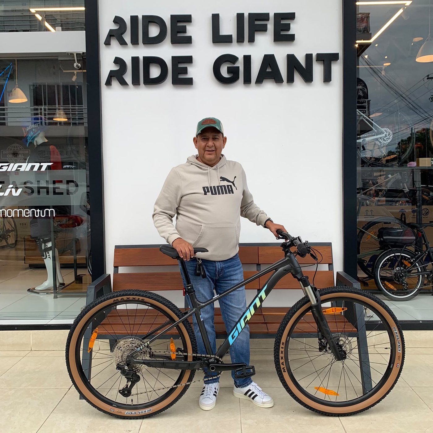 Cliente con su nueva bicicleta de montaña Giant negra de llantas banda café frente al muro Ride Life Ride Giant